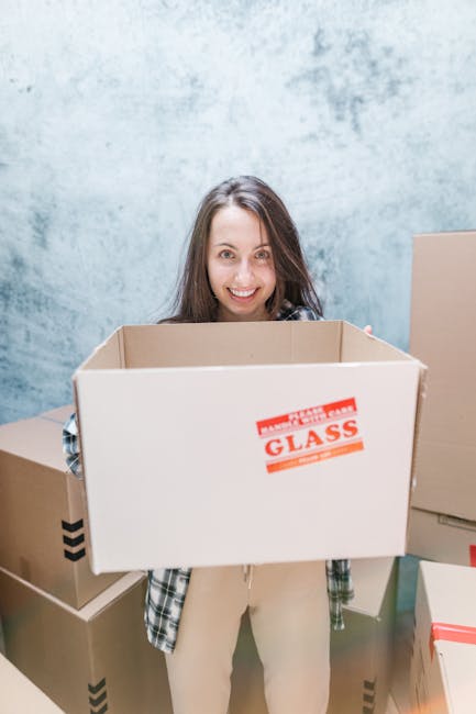 A man and a woman are standing inside a doorway of a residential building, holding cardboard boxes prepared for house relocation. The man, wearing a maroon t-shirt and orange trousers, is holding a la