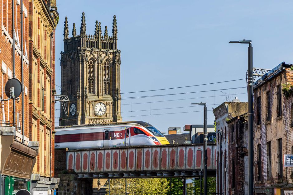 A high-speed train with a streamlined silver, red, and yellow livery is seen crossing an elevated railway bridge in an urban setting, with residential and commercial buildings lining the street below.
